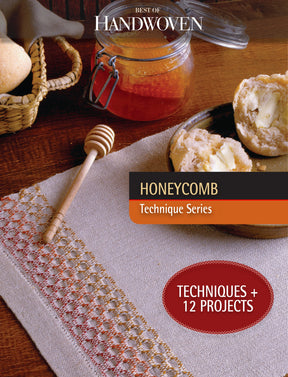 A warm and inviting setting highlights a woven placemat with a honeycomb motif. The table is adorned with a jar of honey, a honey dipper, and a woven basket filled with bread alongside a buttered roll. Text reads: "Best of Handwoven Technique Series: Honeycomb (Printed Version)" by Long Thread Media, featuring Techniques + 12 Projects.