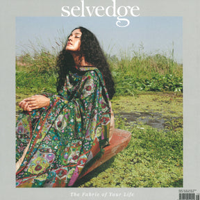 A woman with long, curly hair sits on the edge of a wooden boat surrounded by lush greenery. She is wearing a colorful, patterned dress that reflects exquisite handmade craftsmanship and gazes upwards. The background features tall grasses and a marshy area. The text "Selvedge - Issue 116: Uncut" from the Selvedge brand is at the top of the image.