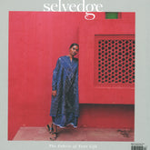 A person in a blue patterned dress poses against a vibrant red wall with intricate latticework, smiling and leaning slightly against the wall. Displayed prominently is the cover of "Selvedge - Issue 117: Irresistible," featuring the tagline "The Fabric of Your Life" and celebrating the calico craze sweeping fashion trends.