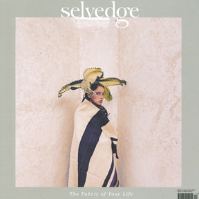 A person wrapped in a striped, woven blanket stands between two beige walls. They are wearing a large, leafy plant as a headpiece, reminiscent of Andy Warhol textile designs. The magazine title "Selvedge - Issue 113: Raw" appears at the top, with the subtitle "The Fabric of Your Life" at the bottom.