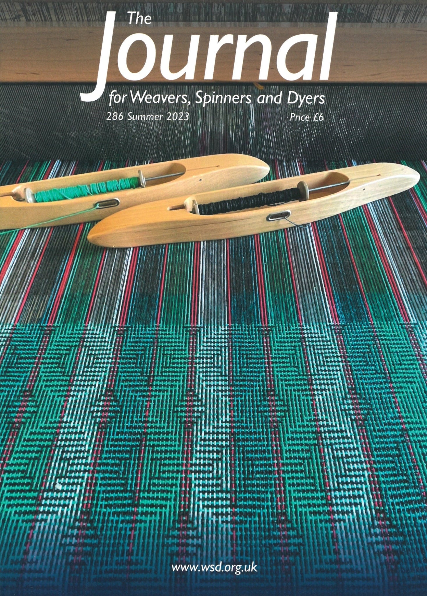 The cover of "The Journal For Weavers, Spinners and Dyers - UK - Issue 286, Summer 2023," priced at £6, showcases two wooden weaving shuttles—one with green thread and one with dark thread—lying on a vividly patterned woven fabric featuring red, green, black, and white designs. This issue includes special insights on botanical printing.