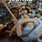 The cover of "The Journal For Weavers, Spinners and Dyers - UK - 287 Autumn 2023" from The Journal For Weavers, Spinners and Dyers features an elegant depiction of a wooden spindle and gray wool roving resting on pebbles, embodying the essence of textile design. A piece of weathered wood serves as the backdrop, with the magazine's URL gracefully displayed below.