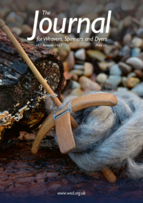 The cover of "The Journal For Weavers, Spinners and Dyers - UK - 287 Autumn 2023" from The Journal For Weavers, Spinners and Dyers features an elegant depiction of a wooden spindle and gray wool roving resting on pebbles, embodying the essence of textile design. A piece of weathered wood serves as the backdrop, with the magazine's URL gracefully displayed below.