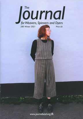 The cover of The Journal for Weavers, Spinners and Dyers UK, Issue 280 (Winter 2021), showcases a person with red hair in a gray and red-striped outfit set against a light purple wall. This edition delves into cloth production and natural dyes. It's available for £6, with the website link included.