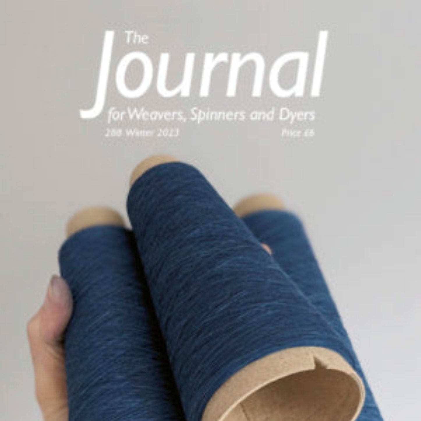 A hand holding three large spools of blue thread, ideal for weaving, set against a light gray background. The top text reads "The Journal for Weavers, Spinners and Dyers, 288 Winter 2023.