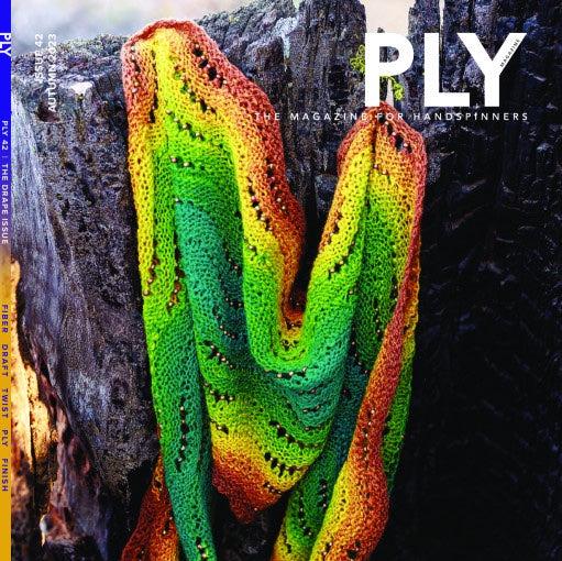 A vibrant knit scarf featuring wavy patterns in green, yellow, red, and brown gracefully rests against a wooden background. Expertly crafted finishing techniques amplify the texture of each fiber. The cover reads, "PLY - The Magazine for Handspinners, Issue 42, Autumn 2023: Drape" from PLY Magazine.