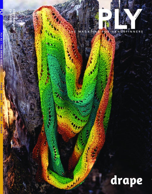 A vibrant knitted scarf in green, orange, and yellow hues is elegantly draped over a log against a rustic, textured backdrop, highlighting its fine finishing techniques. The cover prominently features "PLY Magazine" with the Autumn 2023 issue titled "Drape.