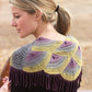 A blonde woman with her hair tied in a ponytail is seen outdoors, standing sideways to the camera, wearing a beautifully knitted shawl featuring a colorful pattern of yellow, purple, and gray semicircles. The shawl comes with fringe and small decorative beads, inspired by the designs found in "Spin-Off Presents: 7 Handspun Shawls to Knit, Crochet, and Weave" by Long Thread Media.