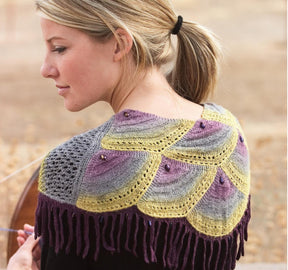 A blonde woman with her hair tied in a ponytail is seen outdoors, standing sideways to the camera, wearing a beautifully knitted shawl featuring a colorful pattern of yellow, purple, and gray semicircles. The shawl comes with fringe and small decorative beads, inspired by the designs found in "Spin-Off Presents: 7 Handspun Shawls to Knit, Crochet, and Weave" by Long Thread Media.