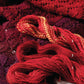 A close-up image showcasing three variations of red and purple yarn, labeled 1, 2, and 3, lying atop a beautifully patterned knitted fabric. The yarns exhibit diverse textures and shades, ranging from vivid red to rich burgundy. This visual is featured in the Spin-Off Presents: "7 Handspun Shawls to Knit, Crochet, and Weave" eBook printed copy by Long Thread Media.