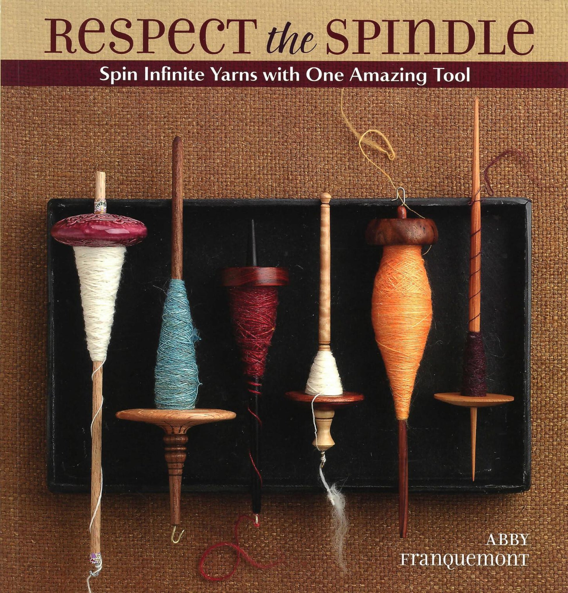 The book cover for "Respect the Spindle" by Abby Franquemont, published by Ingram Content, showcases six unique hand spindles with partially spun yarn in an array of colors against a textured brown fabric background. The subtitle, "Spin Infinite Yarns with One Amazing Tool," invites readers to explore DIY crafts and spinning techniques.