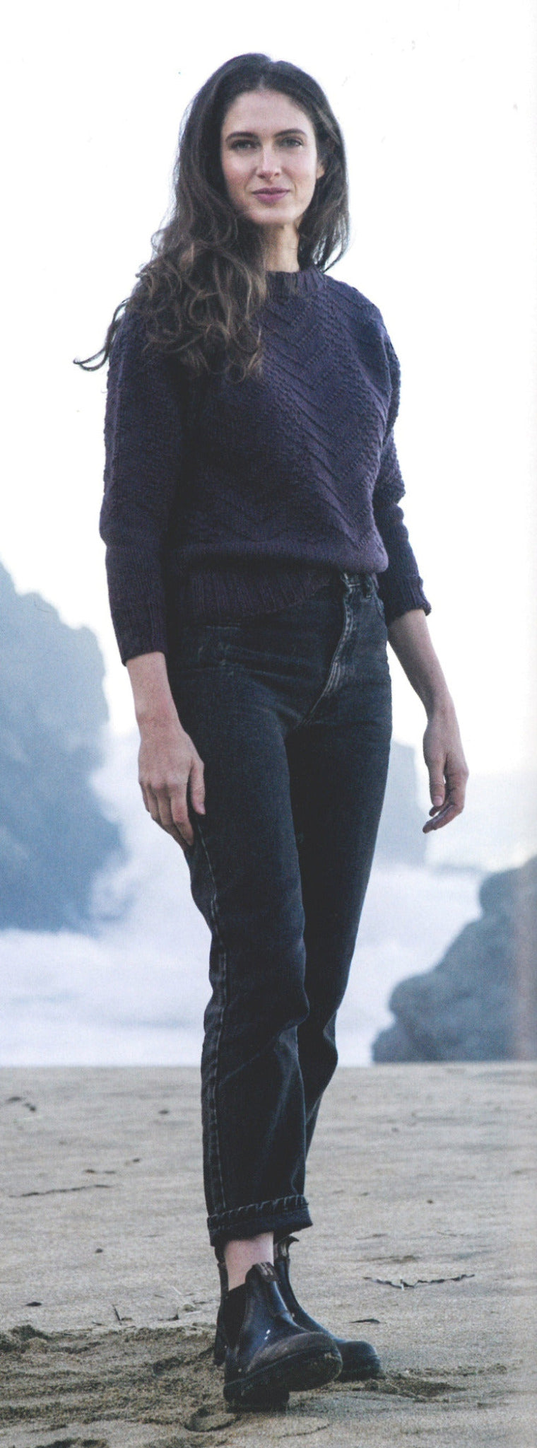 A woman with long wavy hair stands on a foggy beach, wearing a dark sweater reminiscent of those featured in "Knitting Ganseys - Revised and Updated" by Ingram Content. She pairs it with high-waisted black jeans and black boots. The background is filled with mist-covered rocks and waves crashing against the shore as she gazes contentedly towards the camera.