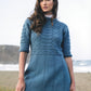 A woman with long hair stands in an outdoor setting near the beach, wearing a blue knitted dress from Ingram Content's "Knitting Ganseys - Revised and Updated," reminiscent of British fisherman's sweaters, featuring intricate patterns. The dress is styled with three-quarter length sleeves and a flared hem. In the background, sand dunes and the ocean are visible.