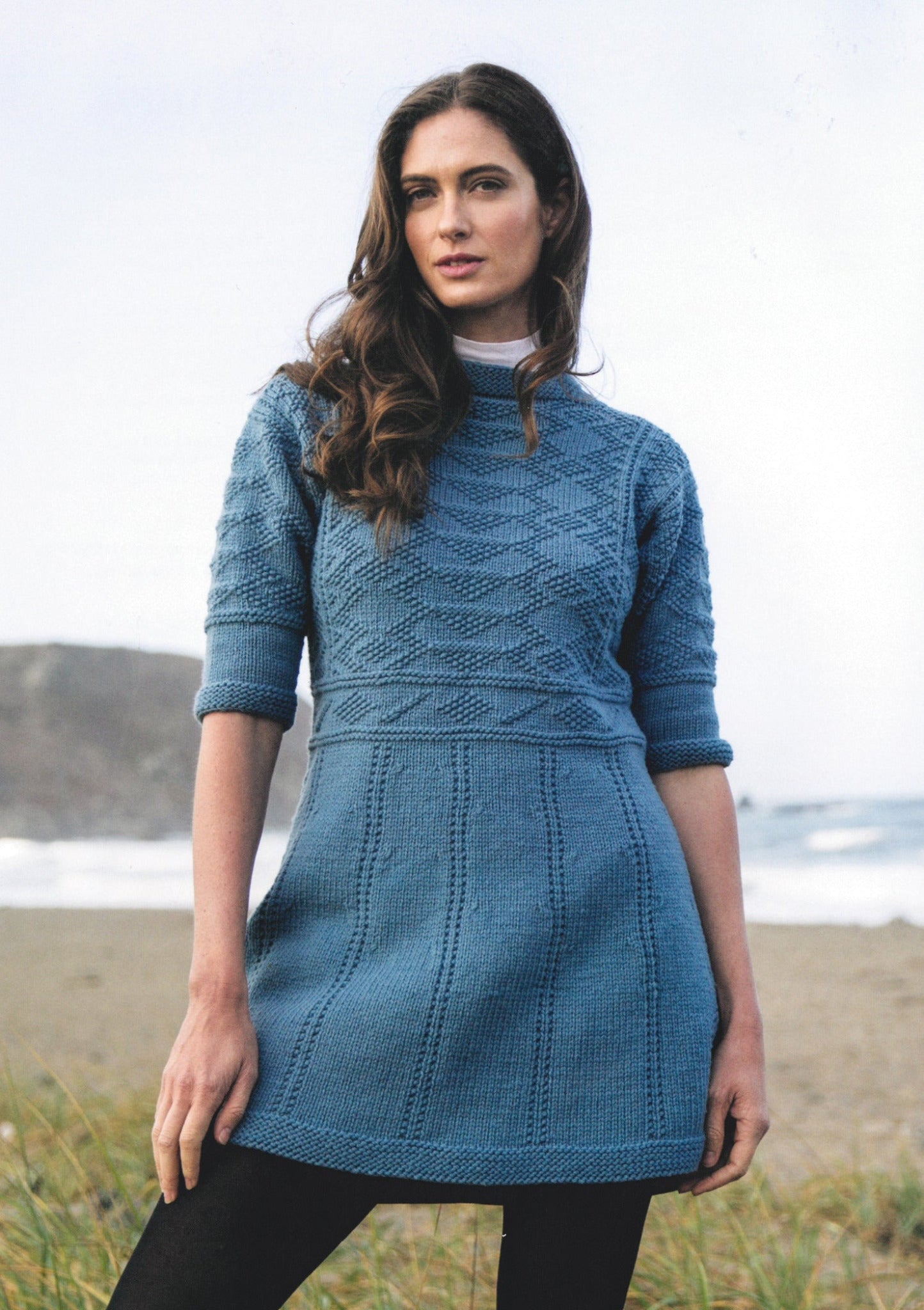 A woman with long hair stands in an outdoor setting near the beach, wearing a blue knitted dress from Ingram Content's "Knitting Ganseys - Revised and Updated," reminiscent of British fisherman's sweaters, featuring intricate patterns. The dress is styled with three-quarter length sleeves and a flared hem. In the background, sand dunes and the ocean are visible.