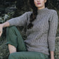 A person with long, braided hair is sitting on a rocky surface, adorned in a cozy gray British fisherman's sweater with a cable pattern from the "Knitting Ganseys - Revised and Updated" collection by Ingram Content. They have rolled-up, olive green pants, and the backdrop features striking textured rock formations. The individual gazes thoughtfully to the right.