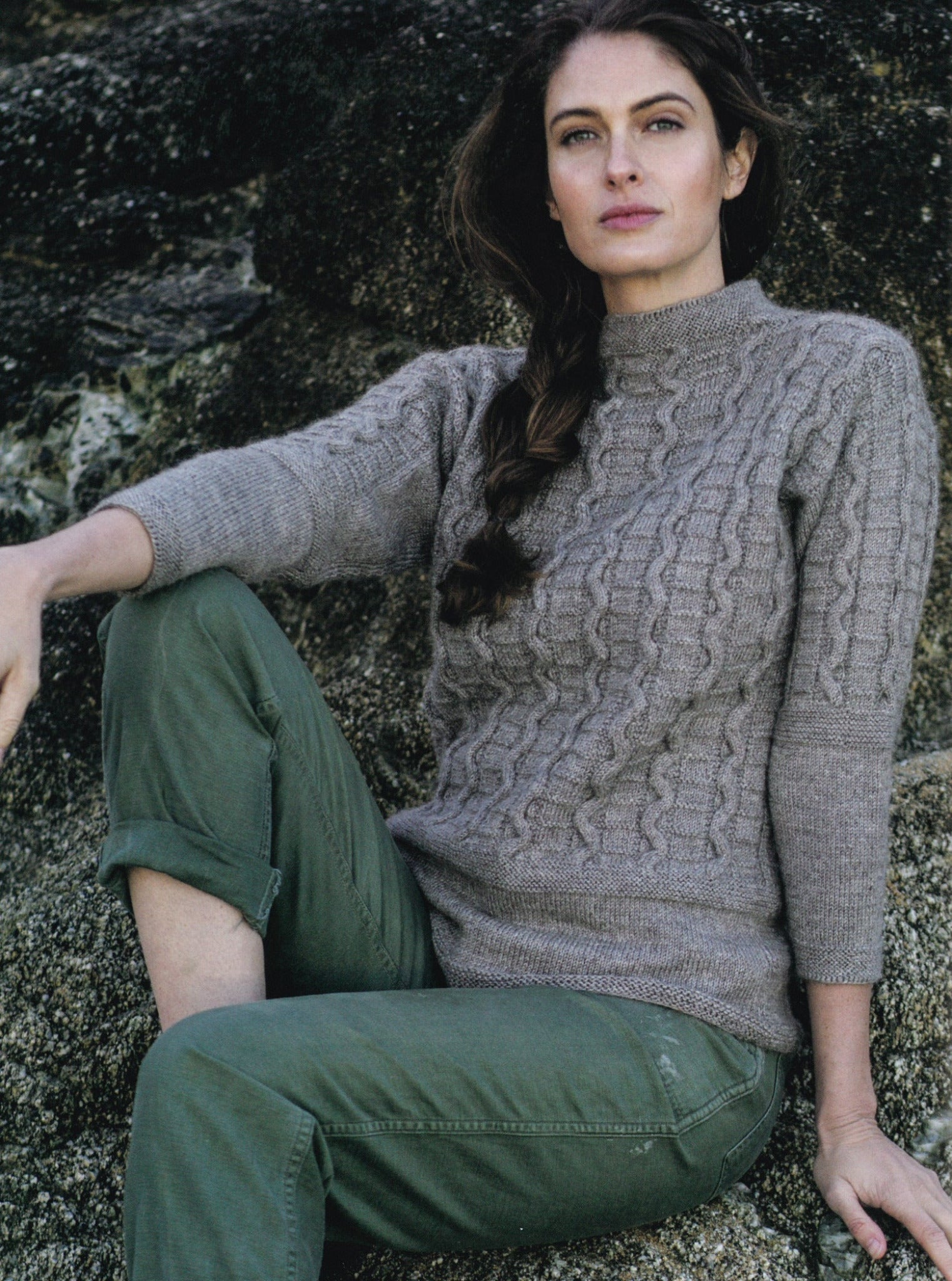 A person with long, braided hair is sitting on a rocky surface, adorned in a cozy gray British fisherman's sweater with a cable pattern from the "Knitting Ganseys - Revised and Updated" collection by Ingram Content. They have rolled-up, olive green pants, and the backdrop features striking textured rock formations. The individual gazes thoughtfully to the right.