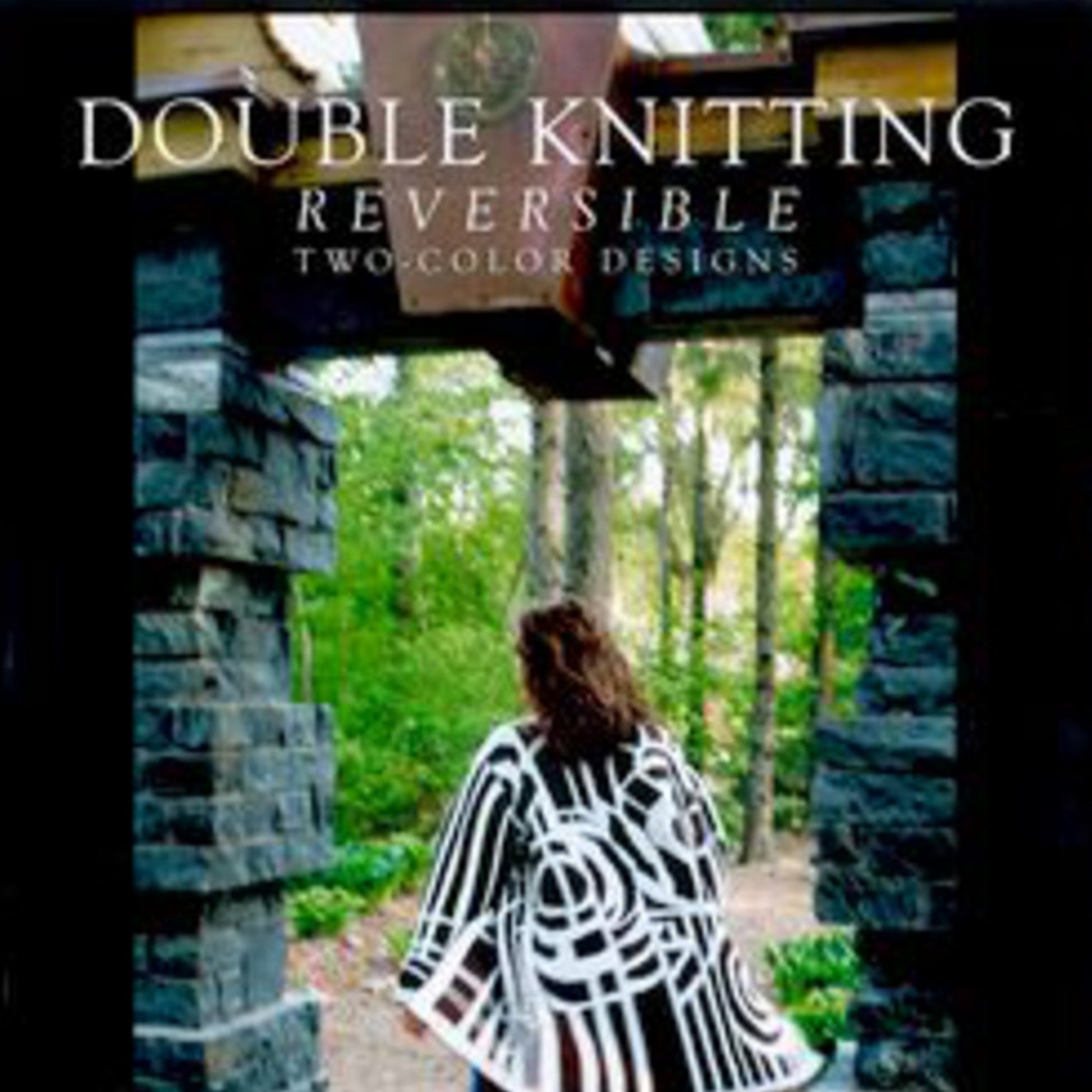 A person with long hair is standing under a stone archway, facing away from the camera, wearing a black and white patterned shawl. Text at the top reads "Double Knitting Reversible Two-Color Designs by Schoolhouse Press." The background shows a lush green garden.