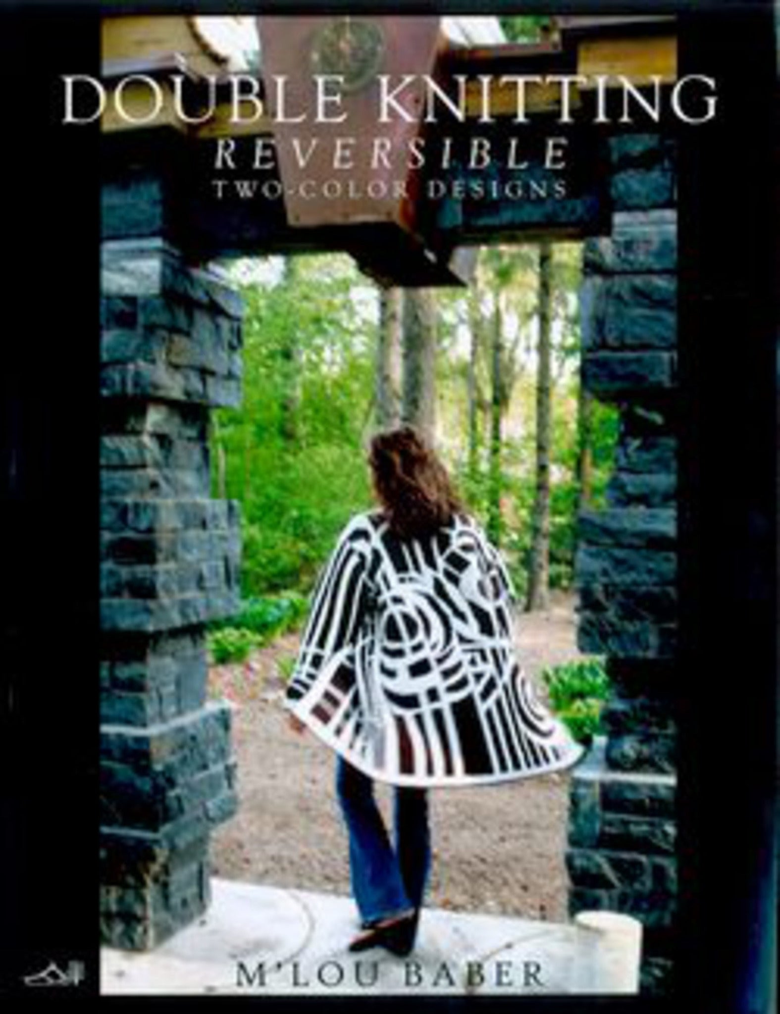 A woman stands between two stone columns, wearing a chic double-knitted reversible cardigan that features stunning abstract patterns from Schoolhouse Press. The background showcases a garden with green trees and a path. The text reads "Double Knitting Reversible Two-Color Designs" by M’Lou Baber.