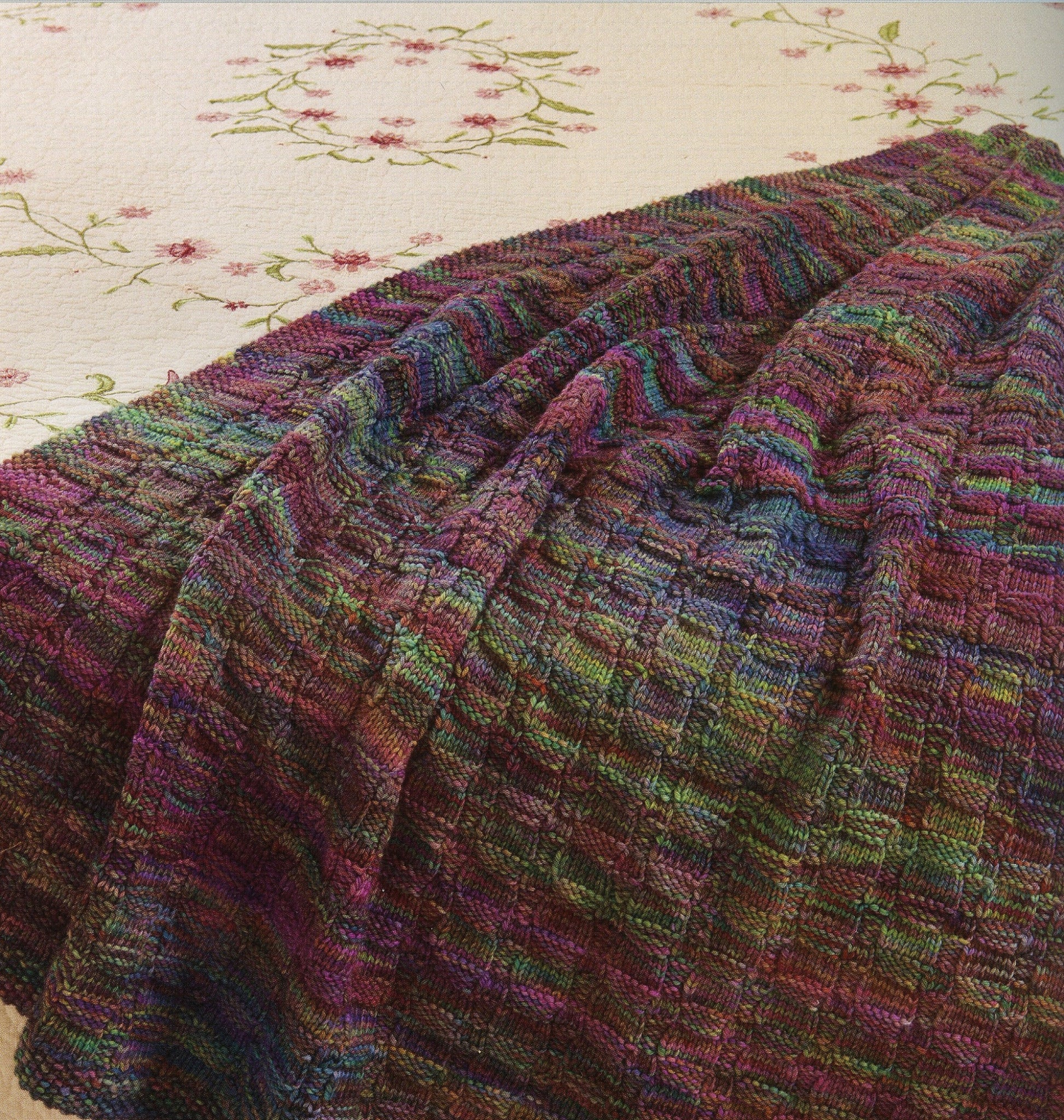 A Pick Your Stitch, Build a Blanket by Martingale & Co, showcasing intricate stitches in a textured, checkered pattern with vibrant multi-colored yarns, is draped over the corner of a floral-patterned, white surface. The vibrant hues of the blanket contrast beautifully with the delicate pink and green floral embroidery beneath it.