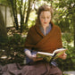 A woman with curly hair wrapped in a brown shawl sits on the grass in a garden, absorbed in reading "The Best of Jane Austen Knits" by Ingram Content. She is surrounded by lush greenery and appears focused on her book. Sunlight filters through the trees, casting dappled light on the scene, echoing the serenity of timeless knitting designs from the Regency era.