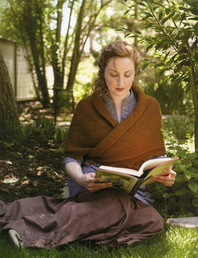 A woman with curly hair wrapped in a brown shawl sits on the grass in a garden, absorbed in reading "The Best of Jane Austen Knits" by Ingram Content. She is surrounded by lush greenery and appears focused on her book. Sunlight filters through the trees, casting dappled light on the scene, echoing the serenity of timeless knitting designs from the Regency era.