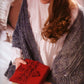 A person wearing a white blouse and a gray knitted shawl is seated, holding "The Best of Jane Austen Knits" book by Ingram Content, which features a red knitted cover. The cozy atmosphere, with a soft-focus background of a pillow and bed, evokes timeless knitting designs reminiscent of Jane Austen's patterns.