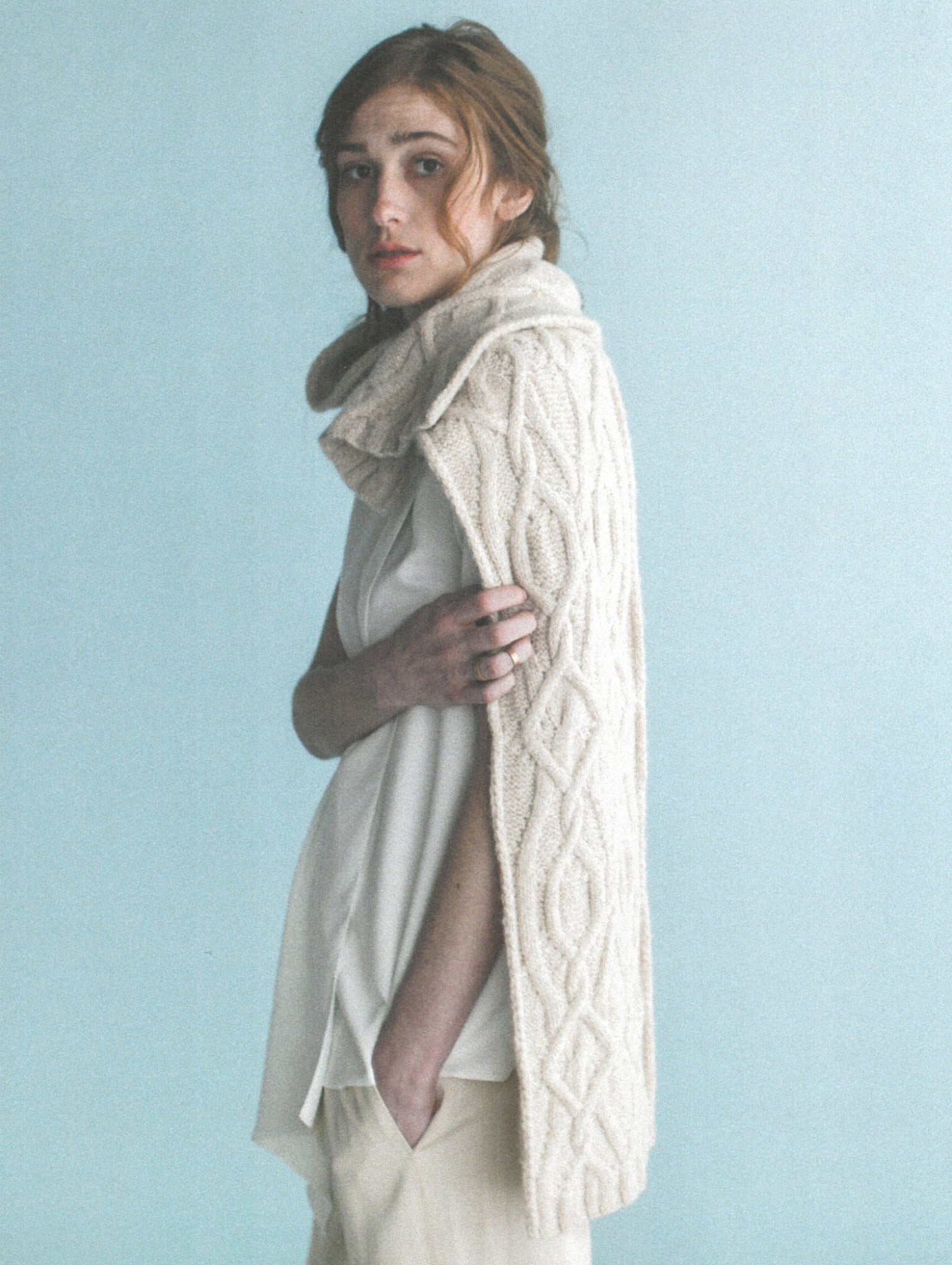 A person with red hair stands against a light blue background, wearing a cream-colored, knitted sleeveless garment from the Norah Gaughan Collection in Wool Studio Vol. 4 by Ingram Content draped over their shoulders and a white blouse. They have a serious expression and their hands are in the pockets of light-colored pants.