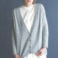 A woman stands against a dark blue background, wearing a light gray, textured, buttoned cardigan over a white, knee-length dress. This beautifully crafted cardigan features exquisite knitting patterns from Wool Studio Vol. 4: The Norah Gaughan Collection by Ingram Content. Her hair is tied up loosely as she looks directly at the camera.