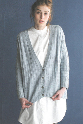 A woman stands against a dark blue background, wearing a light gray, textured, buttoned cardigan over a white, knee-length dress. This beautifully crafted cardigan features exquisite knitting patterns from Wool Studio Vol. 4: The Norah Gaughan Collection by Ingram Content. Her hair is tied up loosely as she looks directly at the camera.