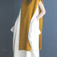 A person with long hair stands against a dark background, wearing a sleeveless mustard-yellow knitted sweater from Ingram Content’s Wool Studio Vol. 4: The Norah Gaughan Collection over a voluminous white dress. The individual has hands in dress pockets and is wearing dark flat shoes.
