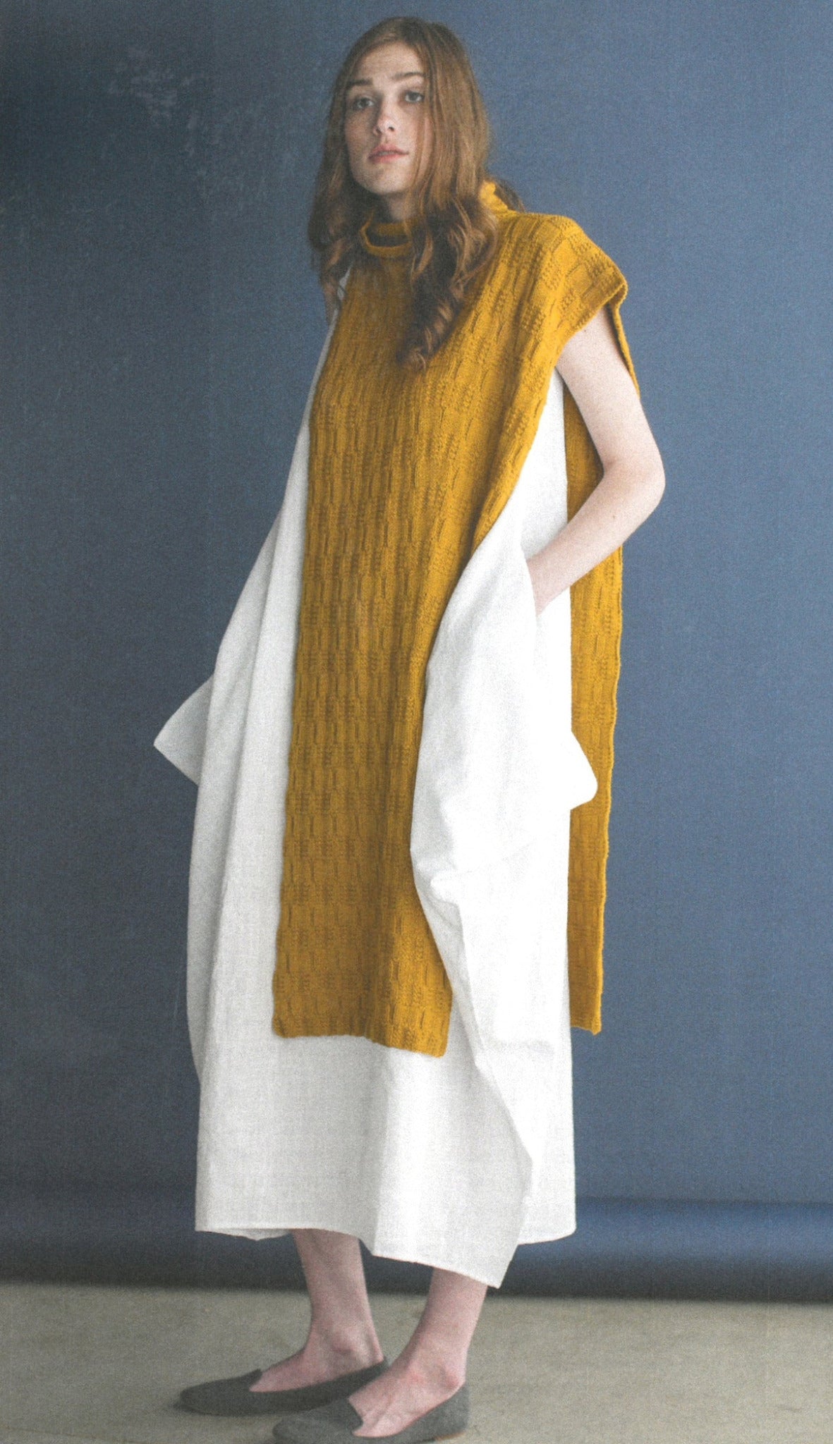 A person with long hair stands against a dark background, wearing a sleeveless mustard-yellow knitted sweater from Ingram Content’s Wool Studio Vol. 4: The Norah Gaughan Collection over a voluminous white dress. The individual has hands in dress pockets and is wearing dark flat shoes.
