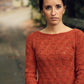 A woman with long hair tied up in a bun stands outdoors, holding a green book. She is wearing an orange sweater from the Botanical Knits collection by Never Not Knitting and large dangling earrings. The background features a blurred path surrounded by lush plant life.