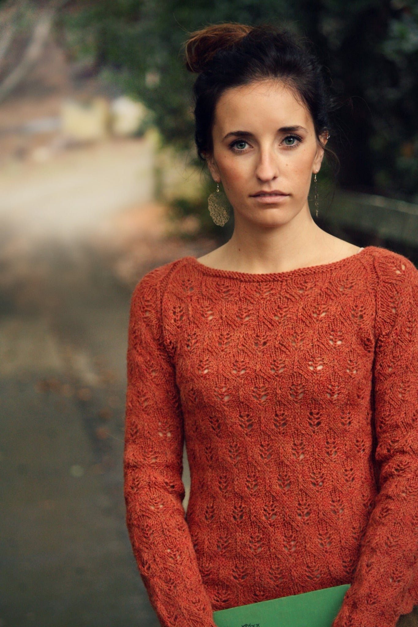 A woman with long hair tied up in a bun stands outdoors, holding a green book. She is wearing an orange sweater from the Botanical Knits collection by Never Not Knitting and large dangling earrings. The background features a blurred path surrounded by lush plant life.