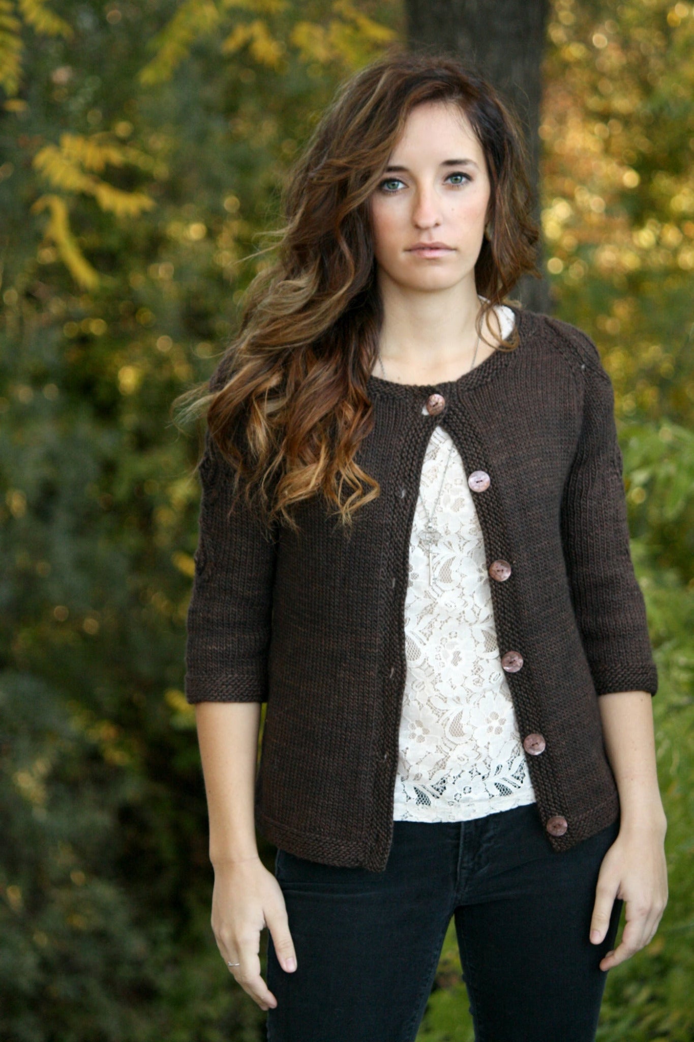 A person with long, wavy hair stands in front of a natural, outdoor background teeming with plant life. They are wearing the "Botanical Knits" brown button-up cardigan from Never Not Knitting over a white lace top and black pants. They are looking directly at the camera with a neutral expression, showcasing their elegant garments effortlessly.