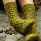 A pair of feet clad in green, knitted Botanical Knits socks by Never Not Knitting, featuring a textured, leaf-like pattern. The feet are resting on a rocky surface, surrounded by a few scattered dried leaves. The socks are thick and cozy, suggesting warmth and comfort—truly elegant garments in the realm of Botanical Knits.