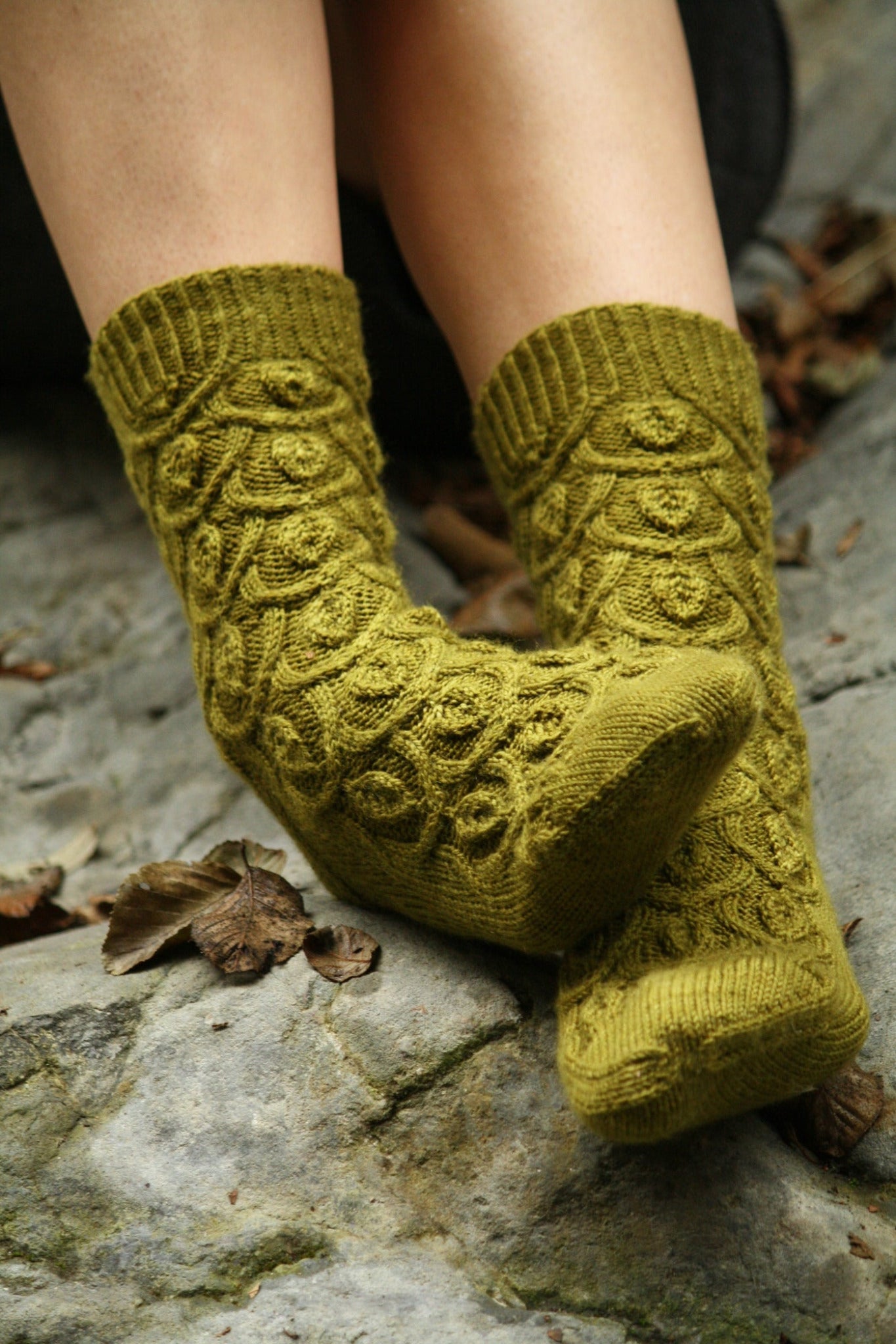 A pair of feet clad in green, knitted Botanical Knits socks by Never Not Knitting, featuring a textured, leaf-like pattern. The feet are resting on a rocky surface, surrounded by a few scattered dried leaves. The socks are thick and cozy, suggesting warmth and comfort—truly elegant garments in the realm of Botanical Knits.