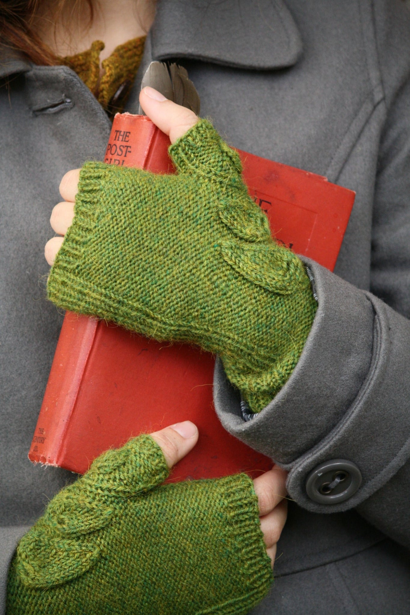 A person dressed in stylish attire, including a gray coat and green fingerless gloves from Never Not Knitting's Botanical Knits collection, is holding a red book. The title of the book, "The Post," is partially visible. The focus remains on the hands, book, and the texture of the clothing.