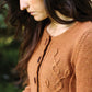 A woman with long, wavy hair looks down thoughtfully. She is wearing a knitted, light brown cardigan inspired by "Botanical Knits 2" from Never Not Knitting, featuring a cable-knit leaf pattern designed by Alana Dakos. The background is slightly blurred, suggesting an outdoor setting.