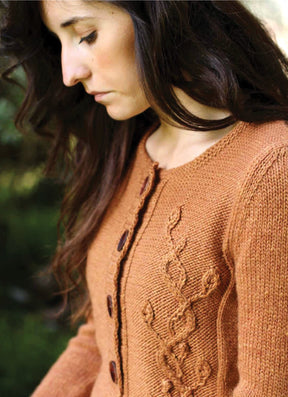 A woman with long, wavy hair looks down thoughtfully. She is wearing a knitted, light brown cardigan inspired by "Botanical Knits 2" from Never Not Knitting, featuring a cable-knit leaf pattern designed by Alana Dakos. The background is slightly blurred, suggesting an outdoor setting.