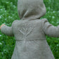 A toddler seen from the back, wearing a knitted, light brown coat with a hood. The coat features a tree design embroidered on the back and is part of our Baby Botanicals collection by Never Not Knitting. The child is standing outdoors on green grass.