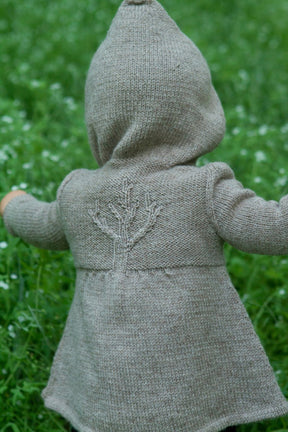 A toddler seen from the back, wearing a knitted, light brown coat with a hood. The coat features a tree design embroidered on the back and is part of our Baby Botanicals collection by Never Not Knitting. The child is standing outdoors on green grass.