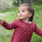 A young girl with short brown hair is wearing a knitted maroon sweater featuring intricate designs from the Baby Botanicals collection by Never Not Knitting, paired with gray pants. She stands in a green, grassy area surrounded by trees and gazes off to the side with a curious expression. One of her arms is slightly extended, embodying the essence of Baby Botanicals.