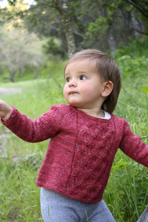 A young girl with short brown hair is wearing a knitted maroon sweater featuring intricate designs from the Baby Botanicals collection by Never Not Knitting, paired with gray pants. She stands in a green, grassy area surrounded by trees and gazes off to the side with a curious expression. One of her arms is slightly extended, embodying the essence of Baby Botanicals.