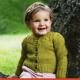 A smiling toddler, adorned in a yellow knitted cardigan from the Never Not Knitting's Baby Botanicals collection, featuring a button-down front, and complemented with a headband. The child is positioned in front of a large tree trunk amidst grassy surroundings, likely a park or forest, with a lush and green backdrop.