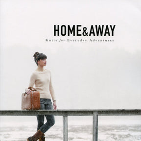 Photo of a book cover titled "Home & Away - Knits for Everyday Adventures" by Hannah Fettig, from Never Not Knitting. It features a woman wearing a knitted sweater, standing on a bridge in a foggy landscape, holding a brown suitcase. The serene and misty background suggests versatile knitting patterns for journey and exploration.