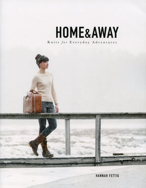 A woman in a cream sweater, jeans, and brown boots stands on a snowy bridge holding a brown suitcase. The cover text reads, "Home & Away - Knits for Everyday Adventures" by Hannah Fettig from Never Not Knitting, featuring versatile knitting patterns. The backdrop is a peaceful winter landscape.