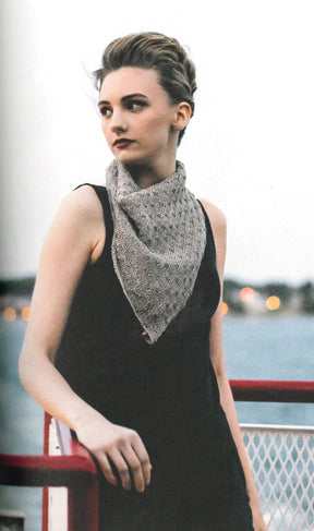 A woman stands outdoors, leaning on a railing with a body of water in the background. She has short, swept-back hair and wears a dark sleeveless top with the "Texture - Exploring Stitch Patterns in Knitwear" by Never Not Knitting—a knitted, triangular scarf around her neck. The background features blurred lights.