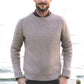 A man with short hair and a beard stands outdoors on a wooden pier by a body of water. He is wearing a beige sweater with intricate stitch patterns from "Texture - Exploring Stitch Patterns in Knitwear" by Never Not Knitting over a maroon shirt and dark pants. Smiling, with his hands in his pockets, the background is slightly out of focus.