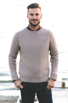 A man with short hair and a beard stands outdoors on a wooden pier by a body of water. He is wearing a beige sweater with intricate stitch patterns from "Texture - Exploring Stitch Patterns in Knitwear" by Never Not Knitting over a maroon shirt and dark pants. Smiling, with his hands in his pockets, the background is slightly out of focus.