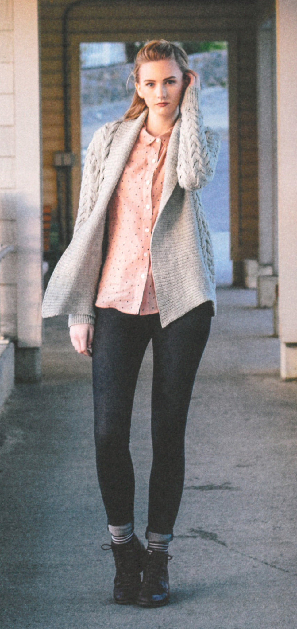 A woman stands outside in a casual outfit. She wears a light pink polka dot shirt, a Never Not Knitting gray knitted cardigan with rich texture from the "Texture - Exploring Stitch Patterns in Knitwear" collection, dark leggings, and dark shoes. She is holding part of her knitwear cardigan with her left hand while standing on a concrete path, with a building in the background.